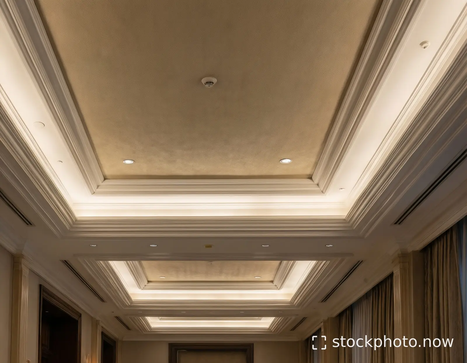 Elegant hotel hallway ceiling with layered crown molding recessed lighting and beige walls.
