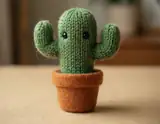 A small green knitted cactus toy with two raised arms and black eyes in an orange felt pot