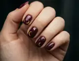 An adult hand with glossy burgundy nails decorated with small gold crescent motifs