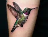 A hyperrealistic tattoo of a hummingbird in mid-flight on a person's thigh with iridescent green and purple feathers.