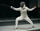 A masked fencer in white protective clothing holding a foil in a wide ready stance
