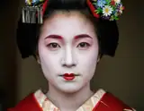 A geisha with white face paint red lips and floral hairpiece gazes forward in traditional red kimono.