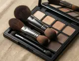 A black eyeshadow palette with brown brushes rests open on a textured beige fabric surface.