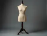 A beige fabric mannequin torso stands on a dark wooden tripod base against a muted gray studio backdrop.