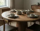 A minimalist wooden dining table set for four with beige ceramic bowls plates and silverware near a window with soft natural light.