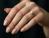 A close-up of a hand with nude manicure wearing a delicate gold ring set with a single lustrous pearl on the ring finger.