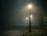 A vintage streetlamp glows warmly through thick fog on a quiet path lined with bare trees and fallen leaves.