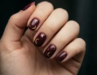 An adult hand with glossy burgundy nails decorated with small gold crescent motifs