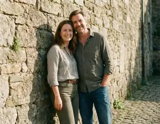 An adult couple standing together beside a textured stone wall in warm sunlight