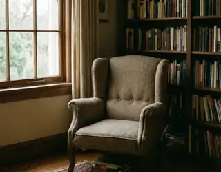 A beige wingback armchair sits by a wooden window beside a tall bookshelf filled with books in a cozy reading nook.