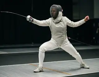 A masked fencer in white protective clothing holding a foil in a wide ready stance