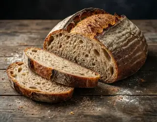 A rustic sourdough loaf with golden crust and airy crumb sits sliced on a flour-dusted wooden table.