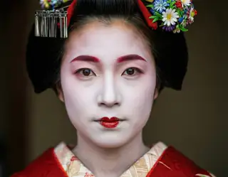 A geisha with white face paint red lips and floral hairpiece gazes forward in traditional red kimono.