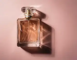 A clear glass perfume bottle with gold cap casts a dramatic shadow on a soft pink background.