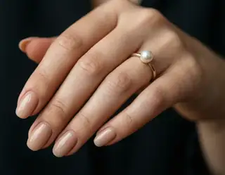 A close-up of a hand with nude manicure wearing a delicate gold ring set with a single lustrous pearl on the ring finger.