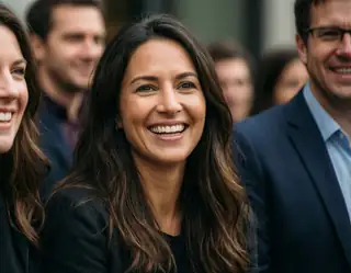 A woman with long dark hair smiles brightly while standing among a group of people in a public setting.