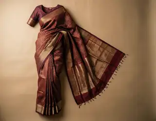 A rich maroon and gold silk saree with intricate woven patterns and tassels draped elegantly against a neutral beige background.