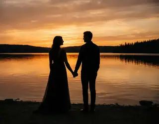 An adult couple holding hands in silhouette beside a calm lake beneath an orange sunset sky