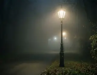 A vintage streetlamp glows warmly through thick fog on a quiet path lined with bare trees and fallen leaves.