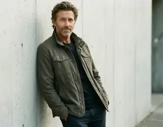 An adult man wearing an olive jacket and leaning beside a pale concrete wall